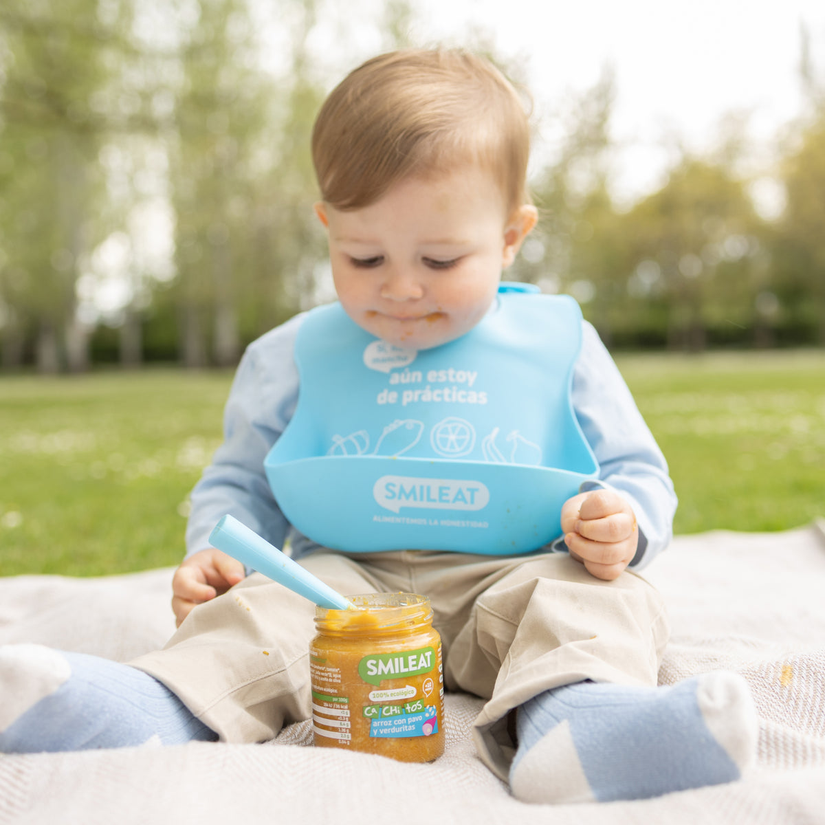 Bebé de babete sentado no parque a comer tarrito biológico – refeição feita com ingredientes reais