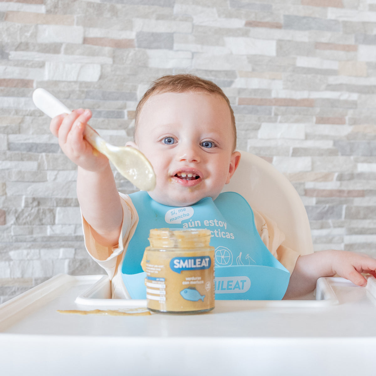 Bebé na cadeira com babete, colher na mão e um boião de comida biológica – refeição com ingredientes reais e honestos